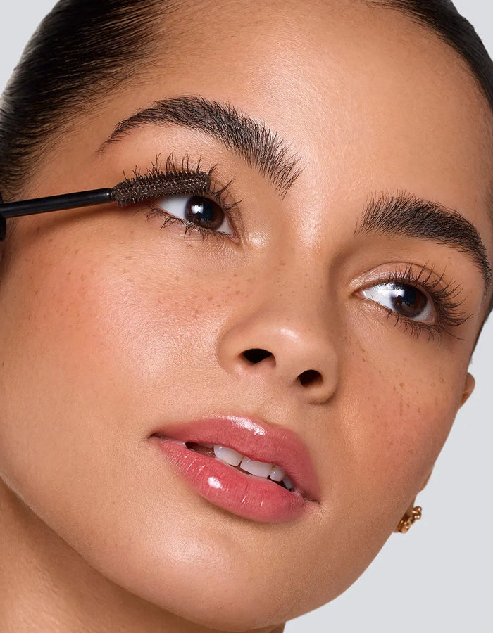 Close-up of a woman applying mascara to her upper lashes with a black wand, showing detailed makeup application on a light gray background.