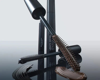 black mascara tubes with applicators and swatches of dark brown mascara, photographed on a reflective dark surface against a light gray background.